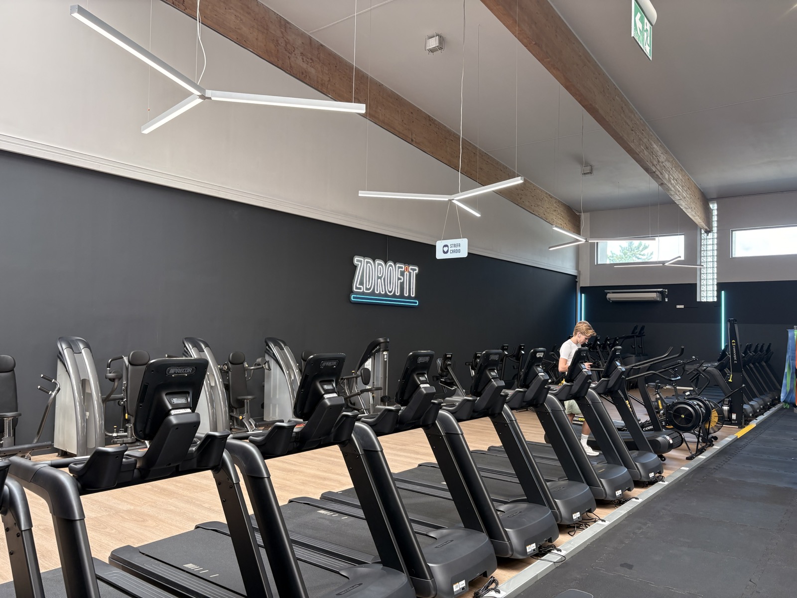 Strefa cardio w Zdrofit Rogowska — rząd bieżni na drewnianej podłodze, neonowe logo Zdrofit na ciemnej ścianie, wioślarze i rower powietrzny