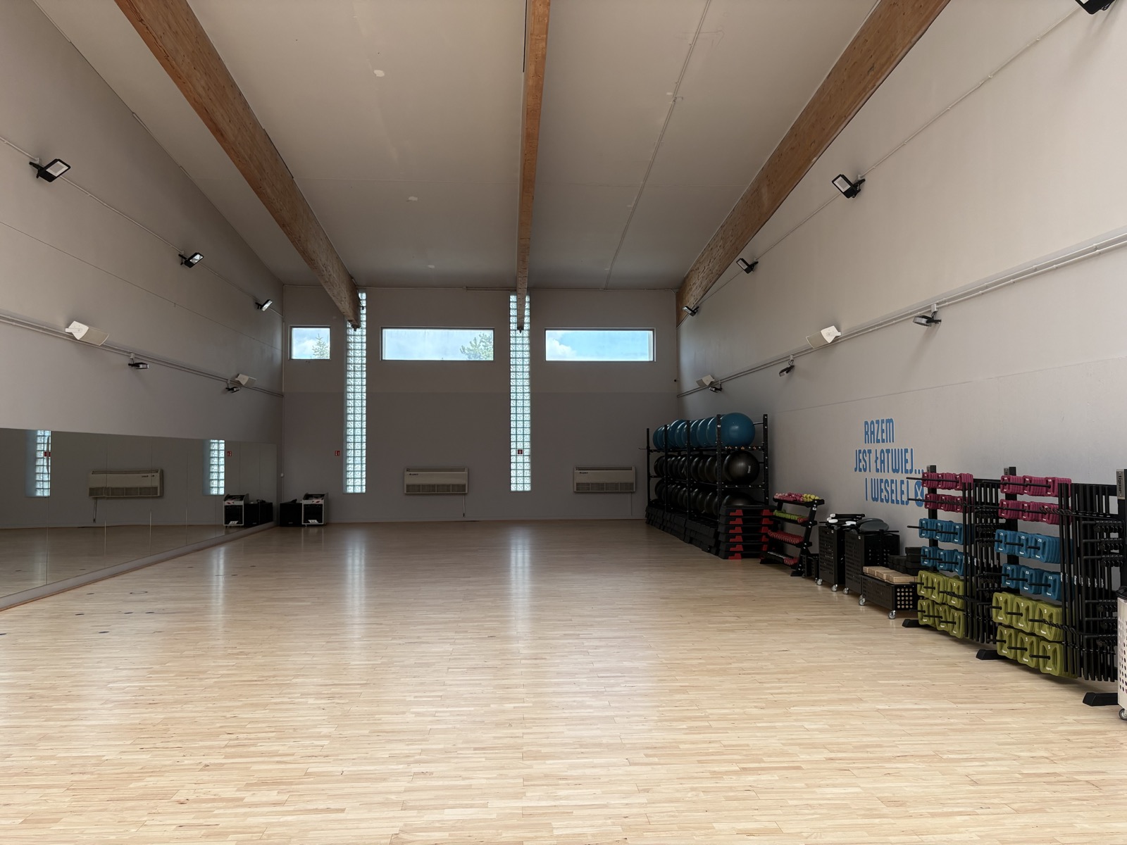 Sala zajęć fitness w Zdrofit Rogowska — parkiet, lustra, piłki, stepy i kolorowe hantle, na ścianie napis Razem jest łatwiej i weselej